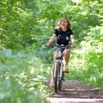 A woman cycling on a scenic forest trail surrounded by green foliage, enjoying a sunny day.